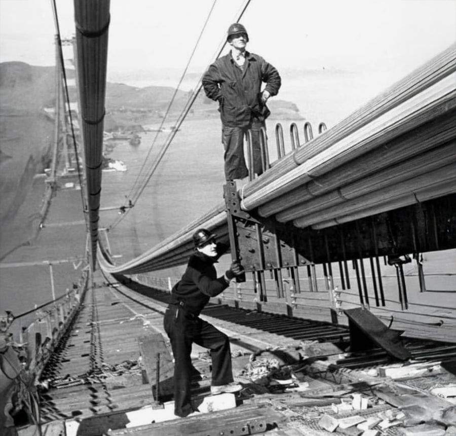Two workers in safety gear repair suspension cables on a bridge, high above the water with land visible in the distance. One stands on the cables, while the other works near steel supports.