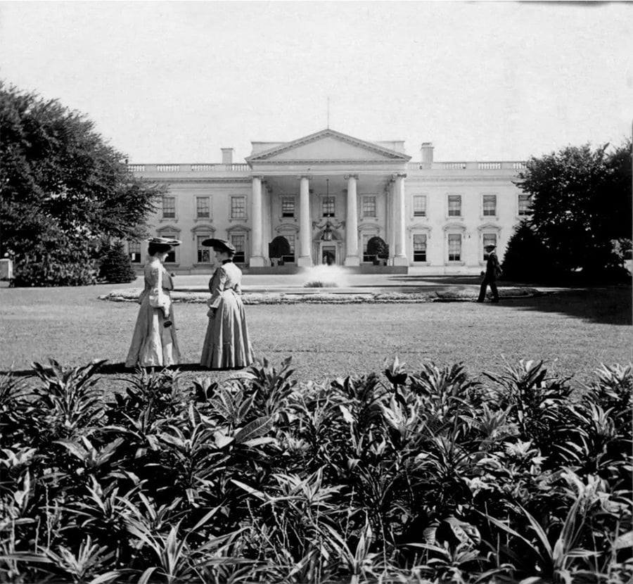Two women in long dresses stand on the lawn in front of the White House, with bushes in the foreground and a person walking in the background, in a black and white photograph.