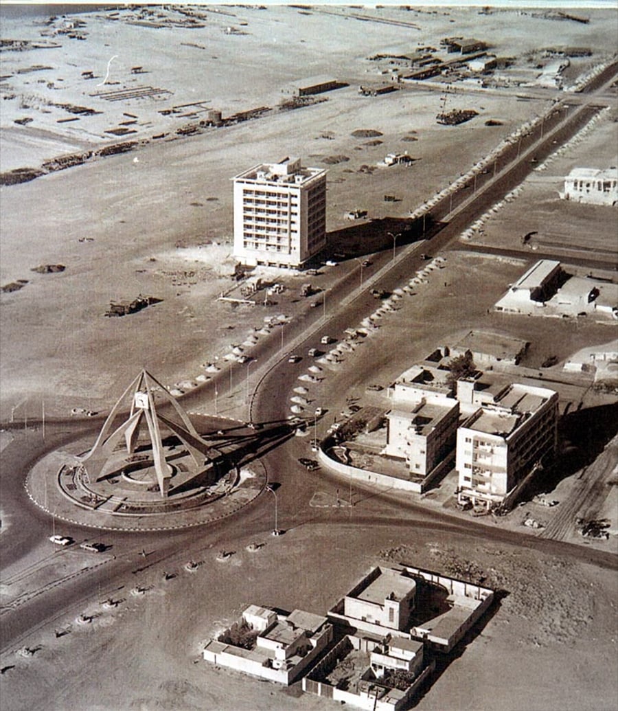 Aerial view of a desert city with a prominent roundabout featuring a large triangular monument, surrounded by low-rise buildings, roads, and scattered vehicles.