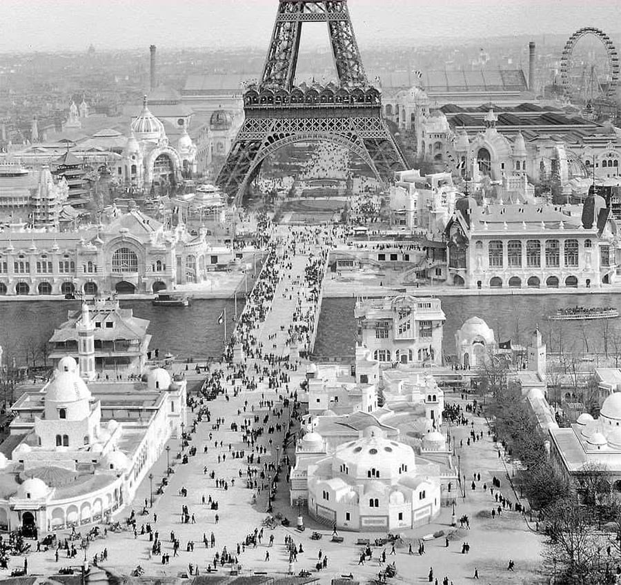 Black and white photo of the Eiffel Tower at the 1900 Paris Exposition, showing crowds walking among ornate exhibition buildings and pavilions near the Seine River.