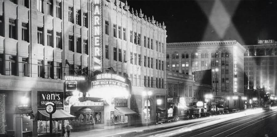 A black-and-white nighttime photo of a city street featuring the illuminated Warner Theatre marquee, nearby storefronts, and blurred car lights indicating motion.