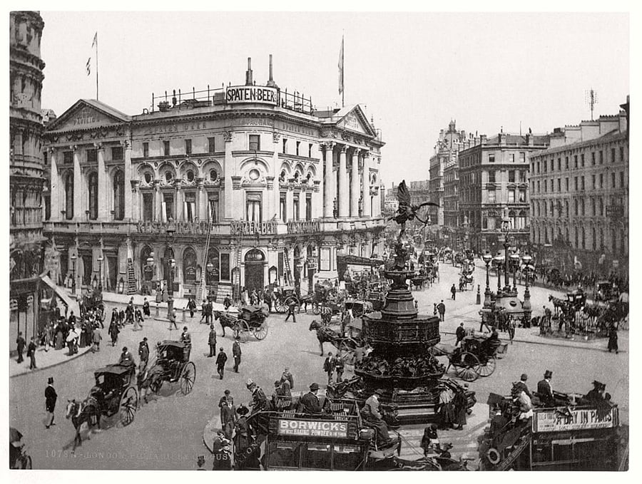 Black-and-white photo of a busy city square with horse-drawn carriages, pedestrians, and ornate buildings. A large decorative fountain stands in the center. Signs for “Spaten Bier” and “Borwick’s” are visible.