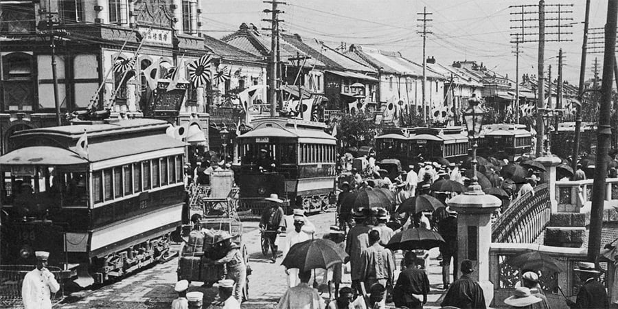 Black-and-white photo of a busy street with trams, power lines, and crowds of people, many holding umbrellas. Traditional buildings line both sides of the street. The scene suggests early 20th-century urban life.