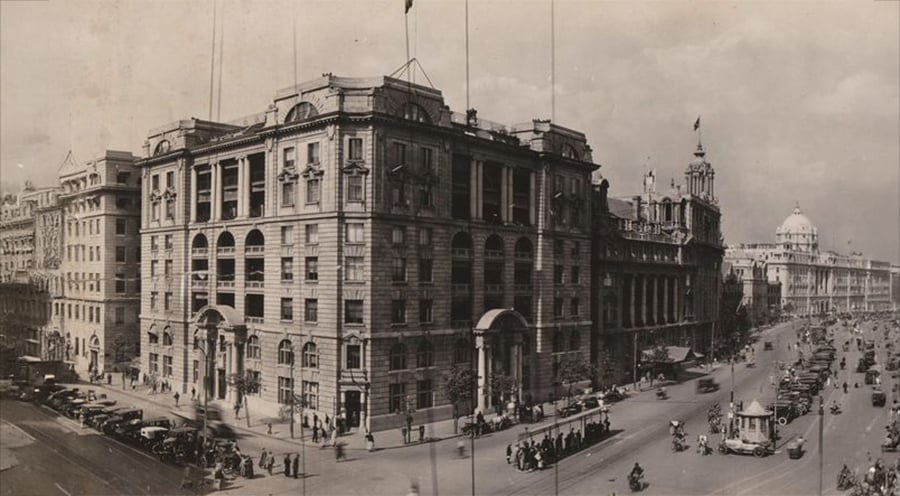 Historic black-and-white photo of a large stone building on a busy street corner, with vintage cars, pedestrians, and streetcars, likely in an early 20th-century city.