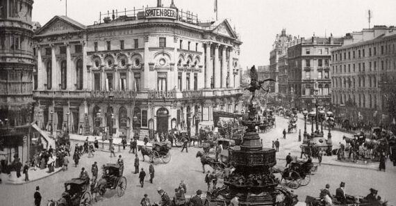 Black and white photo of a busy city square with horse-drawn carriages, people walking, and large historic buildings surrounding a central fountain.
