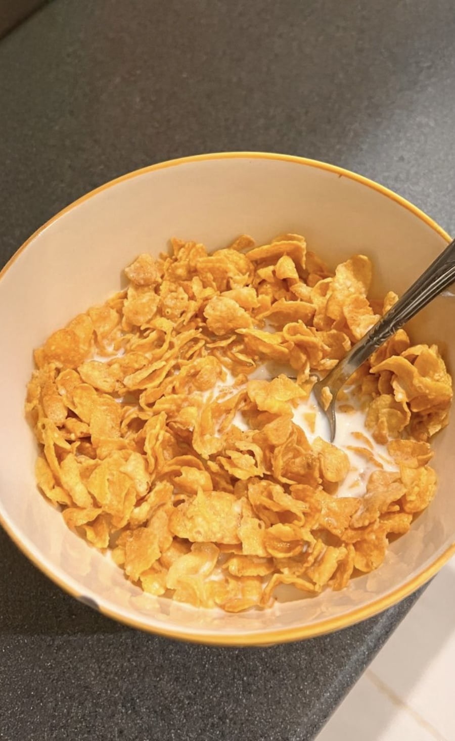 A bowl of cornflakes cereal with milk and a metal spoon on a dark countertop.