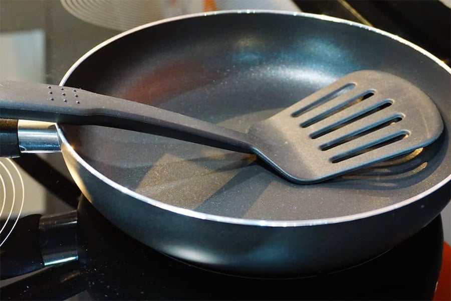 A black frying pan sits on a stovetop with a black slotted spatula resting inside it. The surface of the pan is clean and reflects some light.