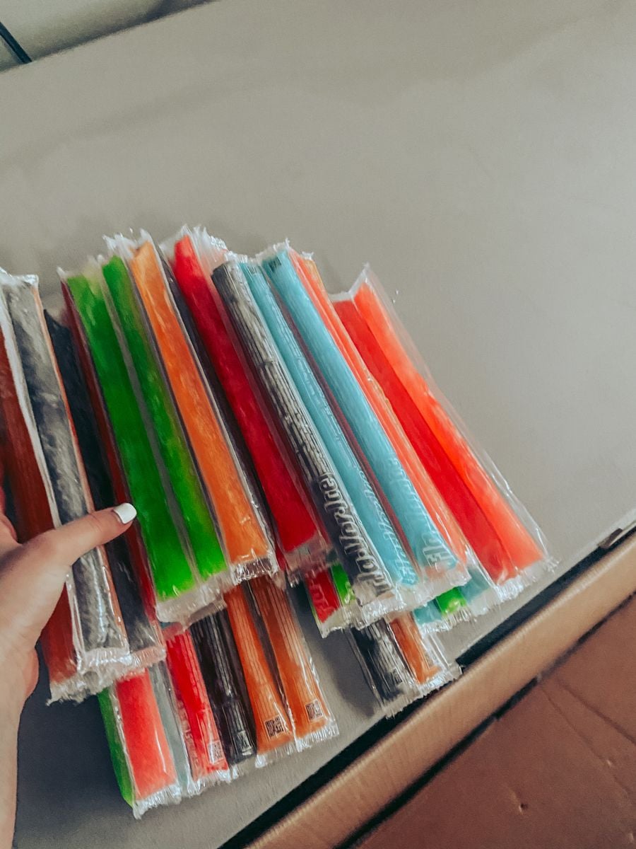 A hand holds a bundle of colorful freeze pops in plastic wrappers, including red, green, orange, blue, and purple, on a beige surface.