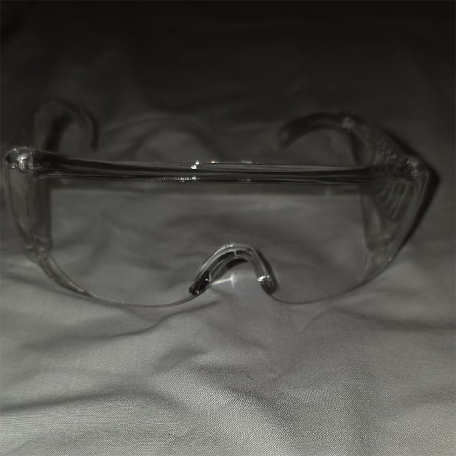 Clear plastic safety glasses resting on a white fabric surface, photographed from the front with light reflection on the lenses.