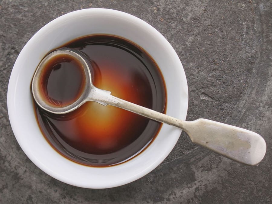 A white bowl filled with dark soy sauce sits on a gray surface. A metal spoon rests in the bowl, partially submerged in the sauce.