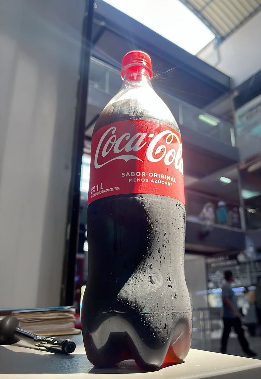 A 1-liter bottle of Coca-Cola with condensation sits on a table indoors. Sunlight shines on the bottle, and a blurred multi-story building is visible in the background.