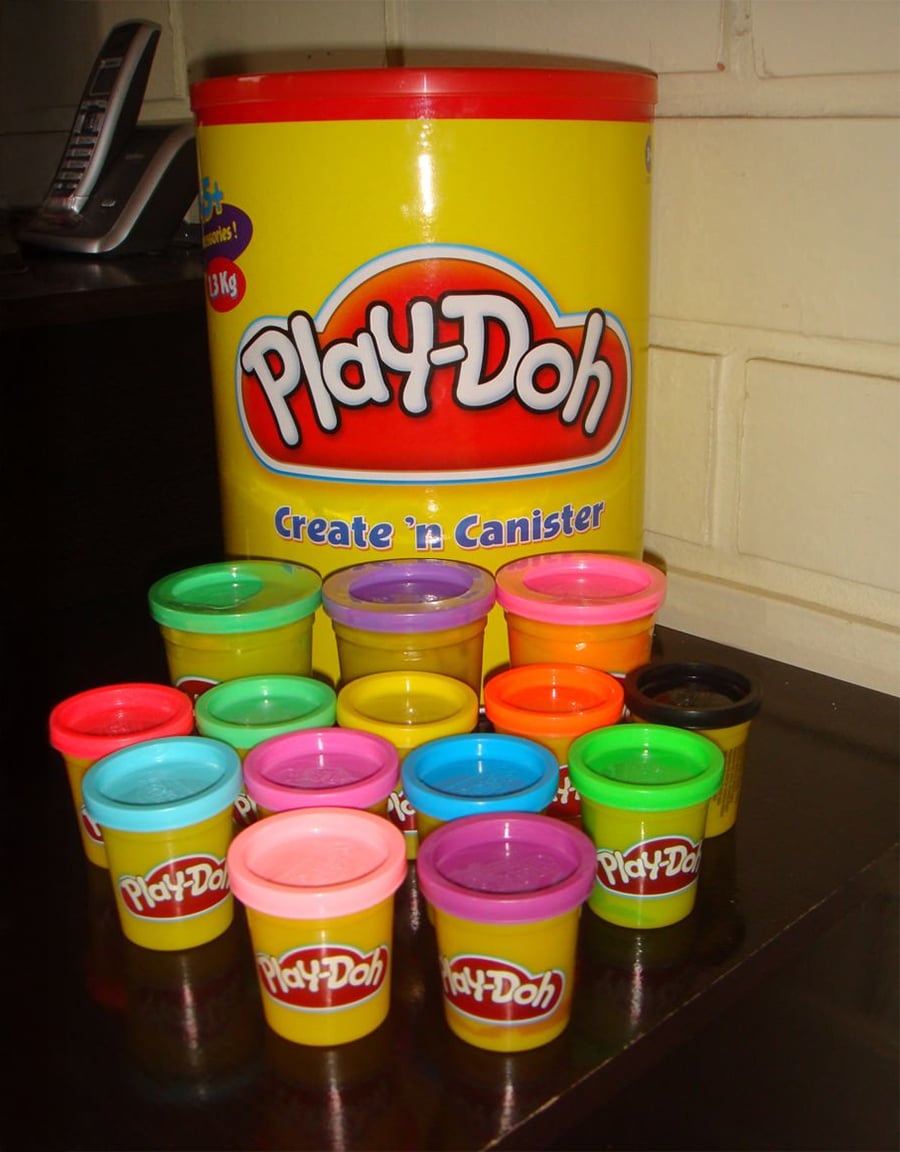 A large yellow Play-Doh canister is behind twelve small cans of Play-Doh in various colors, arranged on a dark surface near a wall and a phone.