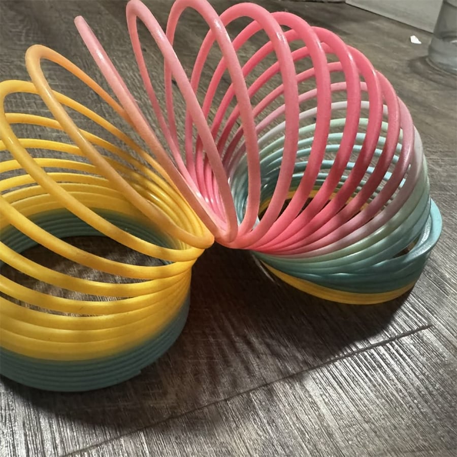 A colorful plastic Slinky toy with yellow, pink, and blue coils is stretched out and sitting on a wooden floor.