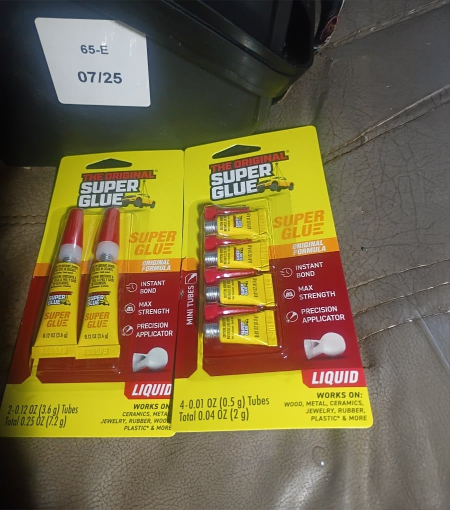 Two yellow-and-red packages of Super Glue containing small tubes are lying on a brown leather surface. One pack has two tubes, the other has four. A white sticker labeled “65-E 07/25” is partially visible above them.