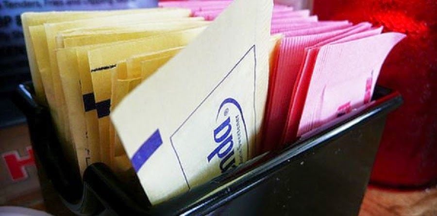 A black container holds packets of yellow, blue, and pink artificial sweeteners, neatly arranged side by side on a table.