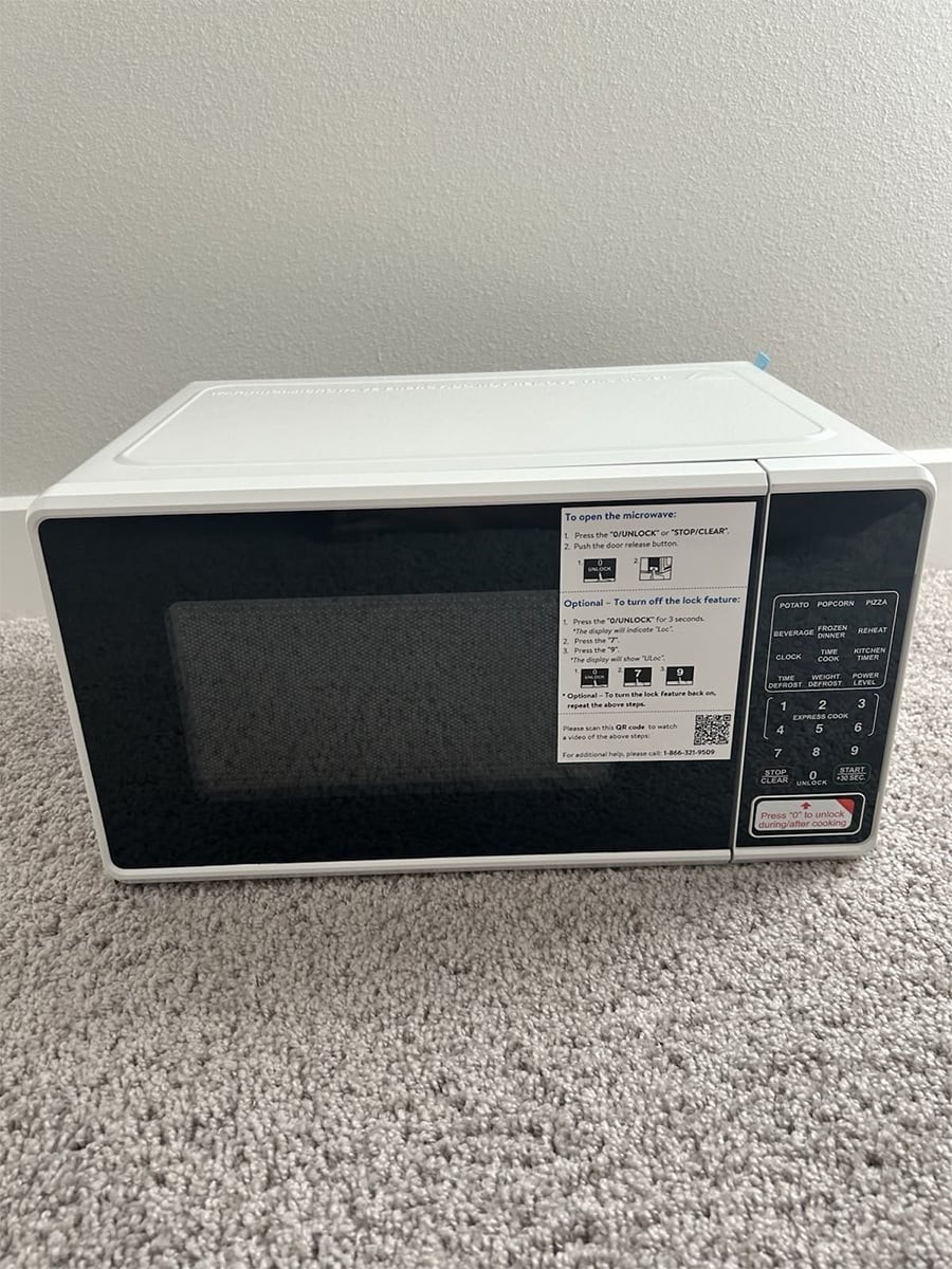 A white microwave with a black front door sits on a beige carpeted floor. There are instructions and buttons visible on the right side of the front panel.