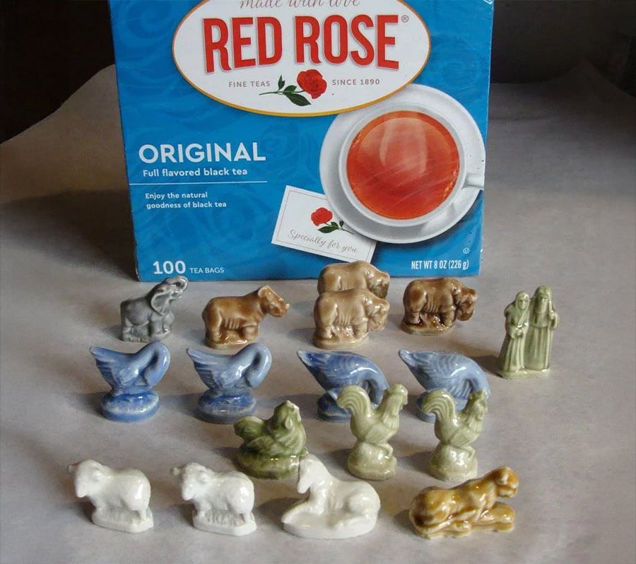 Small ceramic animal figurines and two human figures are arranged in rows in front of a box of Red Rose Original tea bags. The figurines include swans, horses, roosters, and other animals.