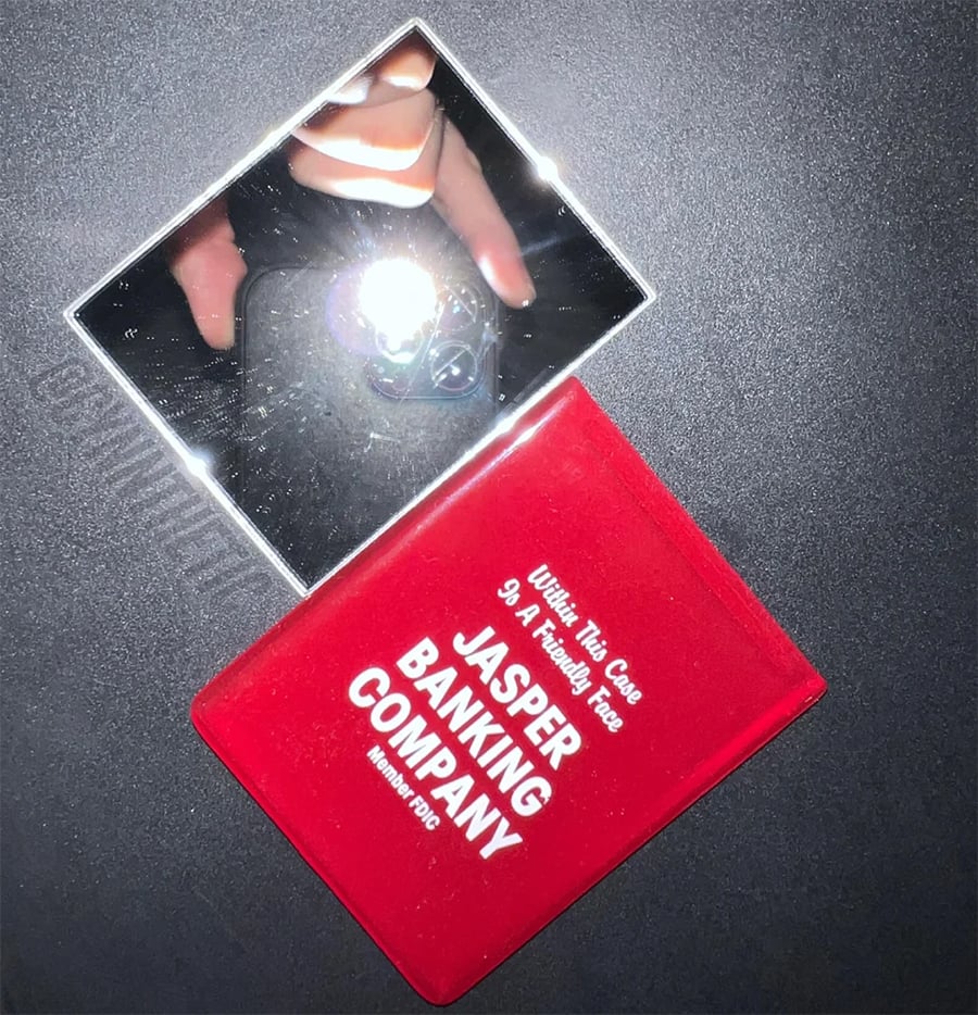 A square mirror reflects a hand holding a phone with a camera flash. Next to it is a red booklet labeled “Jasper Banking Company.” Both items are on a dark, textured surface.