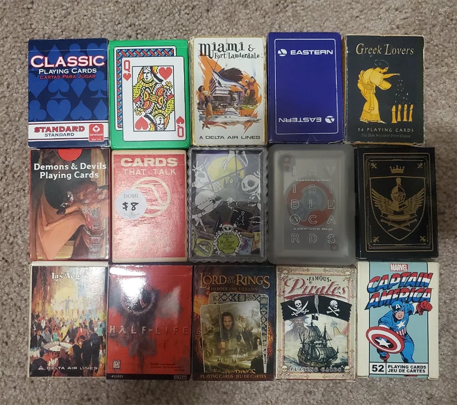 Sixteen different playing card decks arranged in four rows on a beige carpet. Deck themes include airlines, Marvel, pirates, Lord of the Rings, Half-Life, classic, and others, each with unique packaging and artwork.