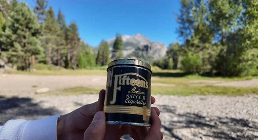 A person holds a can of Jitteen’s Navy Cut Cigarettes outdoors, with trees and distant mountains in the background under a blue sky.