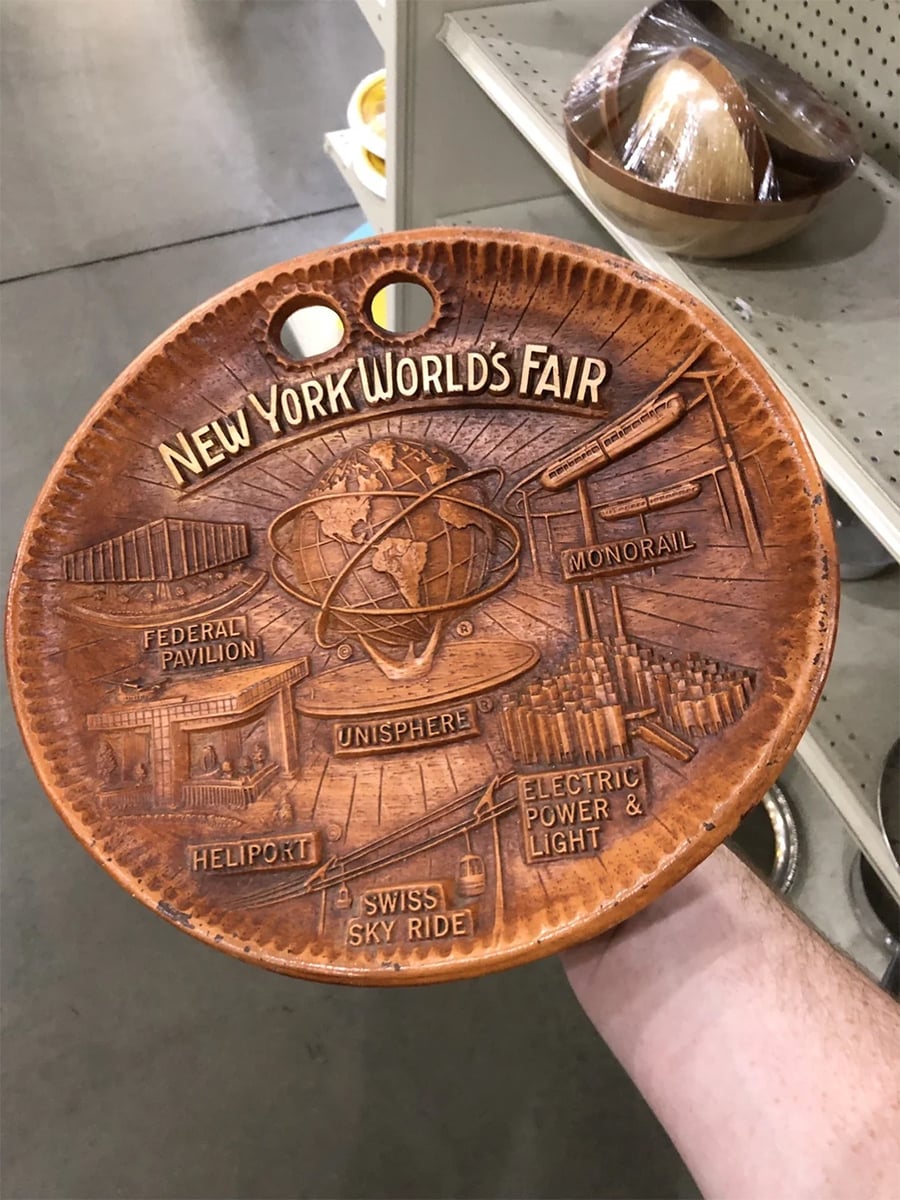 A hand holds a decorative wooden plate featuring raised images and labels from the New York World’s Fair, including Unisphere, Monorail, Heliport, Swiss Sky Ride, Federal Pavilion, and Electric Power & Light.