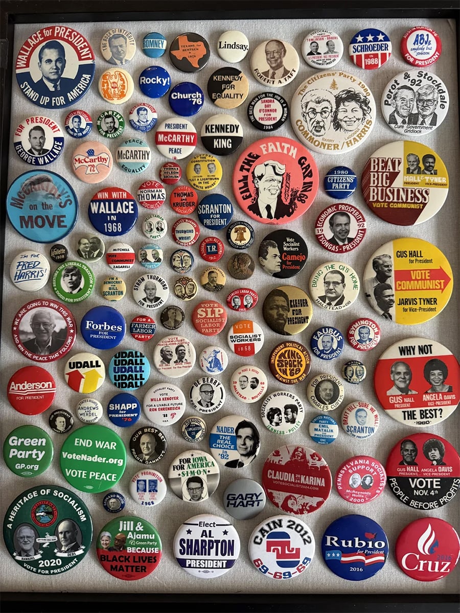 A display of various political campaign buttons from different US elections and eras, featuring multiple candidates, slogans, and party symbols arranged in a grid pattern inside a frame.