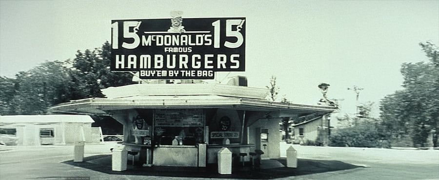 Black and white photo of an old McDonald’s restaurant with a large sign advertising “15¢ McDonald’s Famous Hamburgers.” The building is small with a walk-up counter and is surrounded by trees and a parking lot.