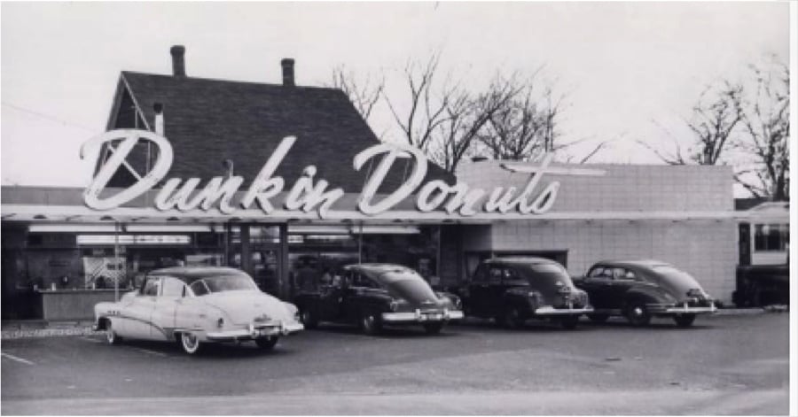 Black-and-white photo of a vintage Dunkin’ Donuts storefront with large cursive sign and four classic cars parked in front. Leafless trees and a house are visible in the background.