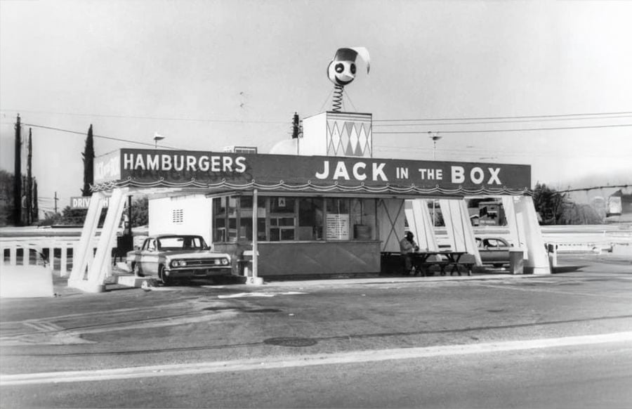 Black-and-white photo of an old Jack in the Box fast-food restaurant with a large clown head on the roof, cars parked out front, and a person sitting at an outdoor table.