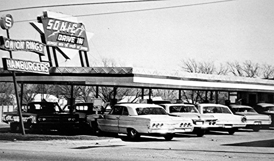 Black and white photo of a vintage drive-in restaurant with classic cars parked under a canopy. A sign reads “Sonic Drive In, Onion Rings, Hamburgers.” Leafless trees are visible in the background.