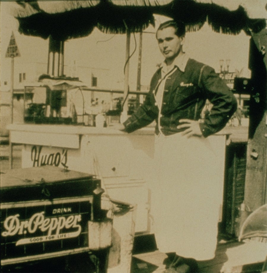 A vintage photo of a man in a uniform and apron standing by a food or drink stand with a sign reading “Drink Dr. Pepper” in the foreground. The scene appears to be from the early 20th century.