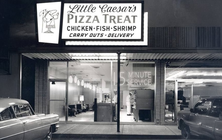 Black and white photo of a vintage Little Caesar’s Pizza Treat restaurant with a lit sign advertising pizza, chicken, fish, shrimp, carry outs, delivery, and 15 minute service; old cars are parked outside.