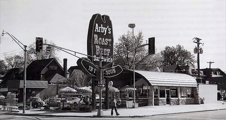 Black and white photo of a vintage Arby’s restaurant with a large cowboy hat sign reading “Arby’s Roast Beef Sandwich” and cars parked outside. A person is walking near the entrance.