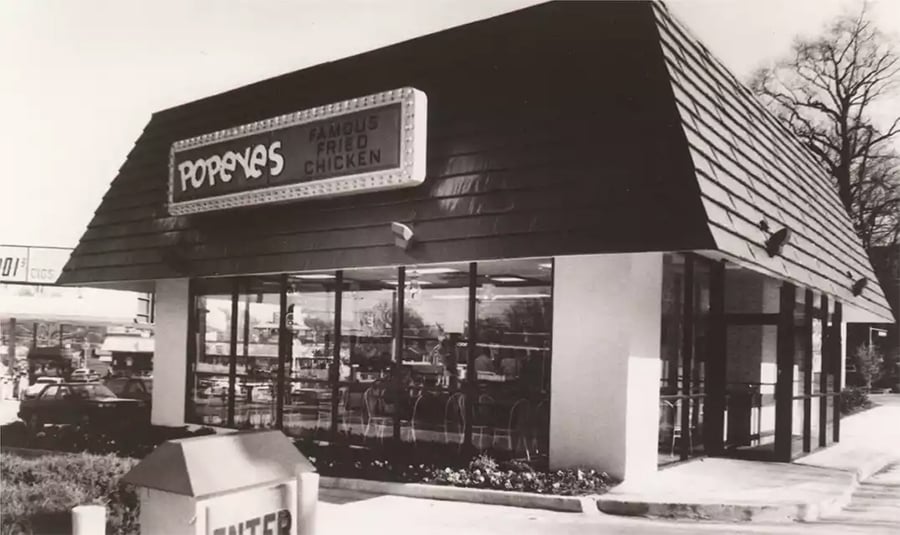 A vintage black-and-white photo of a Popeyes restaurant with a retro sign reading “Popeyes Famous Fried Chicken.” The building has large windows and a dark slanted roof. Cars and trees are visible in the background.