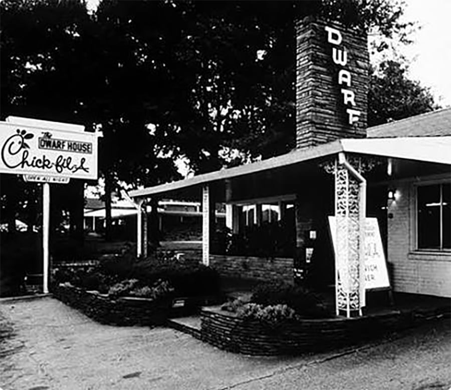 Black and white photo of the original Chick-fil-A restaurant, The Dwarf House, with vintage signs and a covered entrance, surrounded by trees and shrubs.