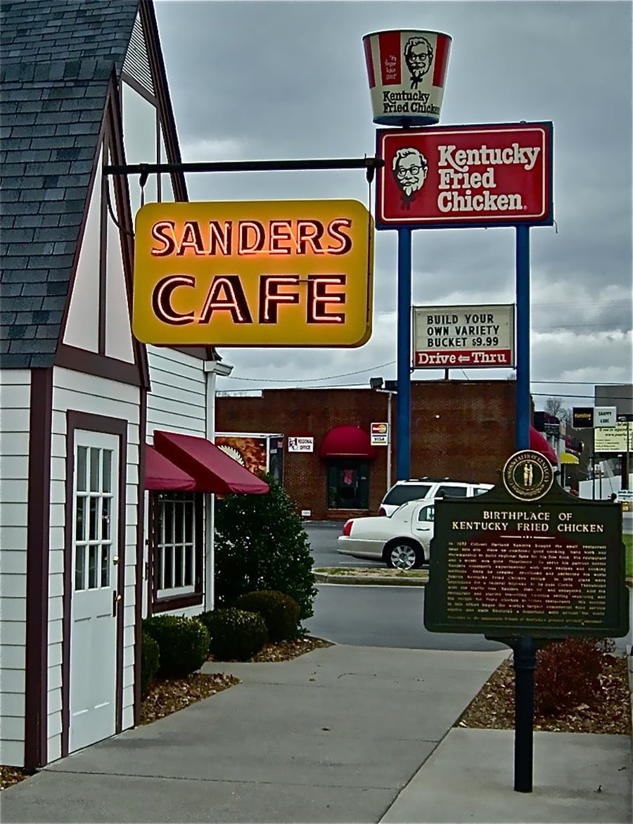 Sanders Cafe and Kentucky Fried Chicken signs stand near a historical marker, marking the birthplace of KFC. A small white building and a cloudy sky are also visible.