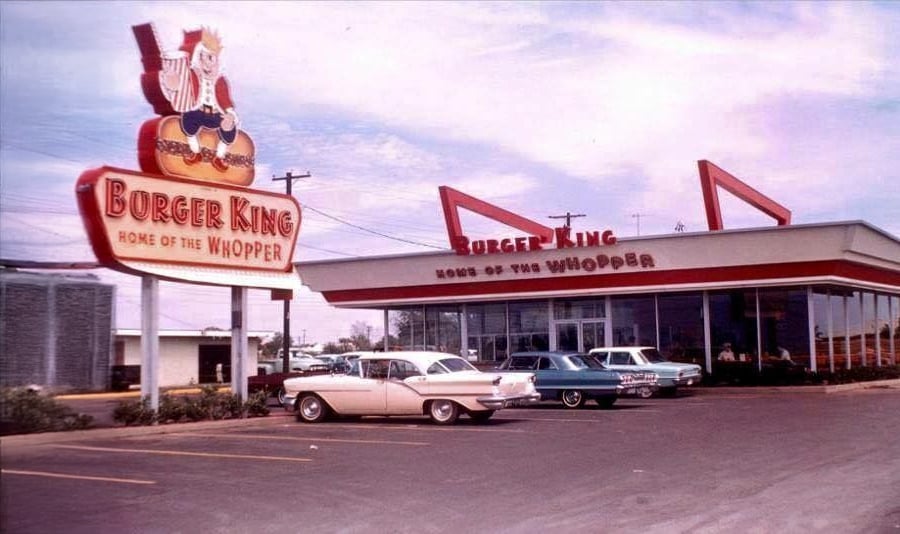 A vintage Burger King restaurant with a large sign featuring the Burger King mascot and two classic cars parked outside. The building and cars suggest the image is from the 1950s or 1960s.