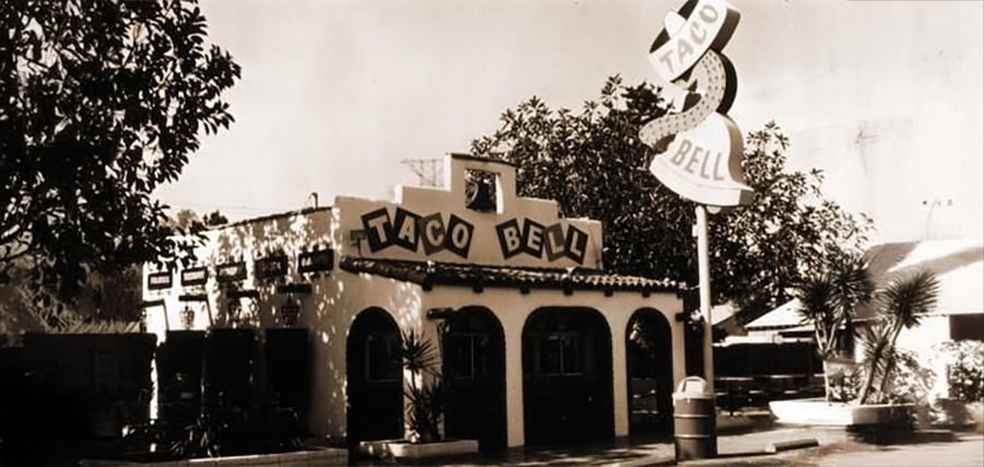A vintage black-and-white photo of the original Taco Bell restaurant with a large retro sign and a small building featuring arched doorways and a tiled roof surrounded by trees.