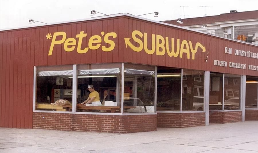 A red building with large yellow letters reading “*Pete’s SUBWAY*.” Inside, a person prepares food behind a counter. The windows reflect the street and surrounding buildings.