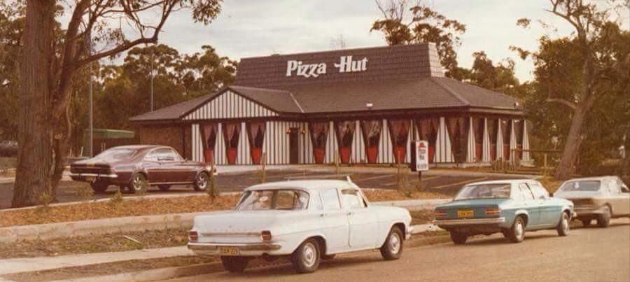 A vintage Pizza Hut restaurant with striped exterior, parked classic cars in front, and trees in the background on a cloudy day.