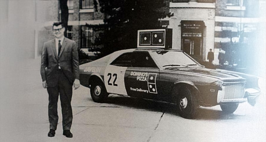 A man in a suit stands next to a vintage Domino’s Pizza delivery car with the number 22 on the door, parked on a city street in front of a brick building.
