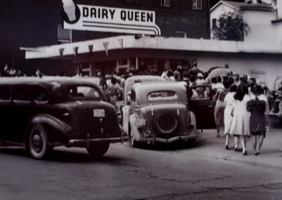 Black and white photo of a crowded Dairy Queen, with people standing outside and vintage cars parked in front; several women in dresses walk towards the shop.