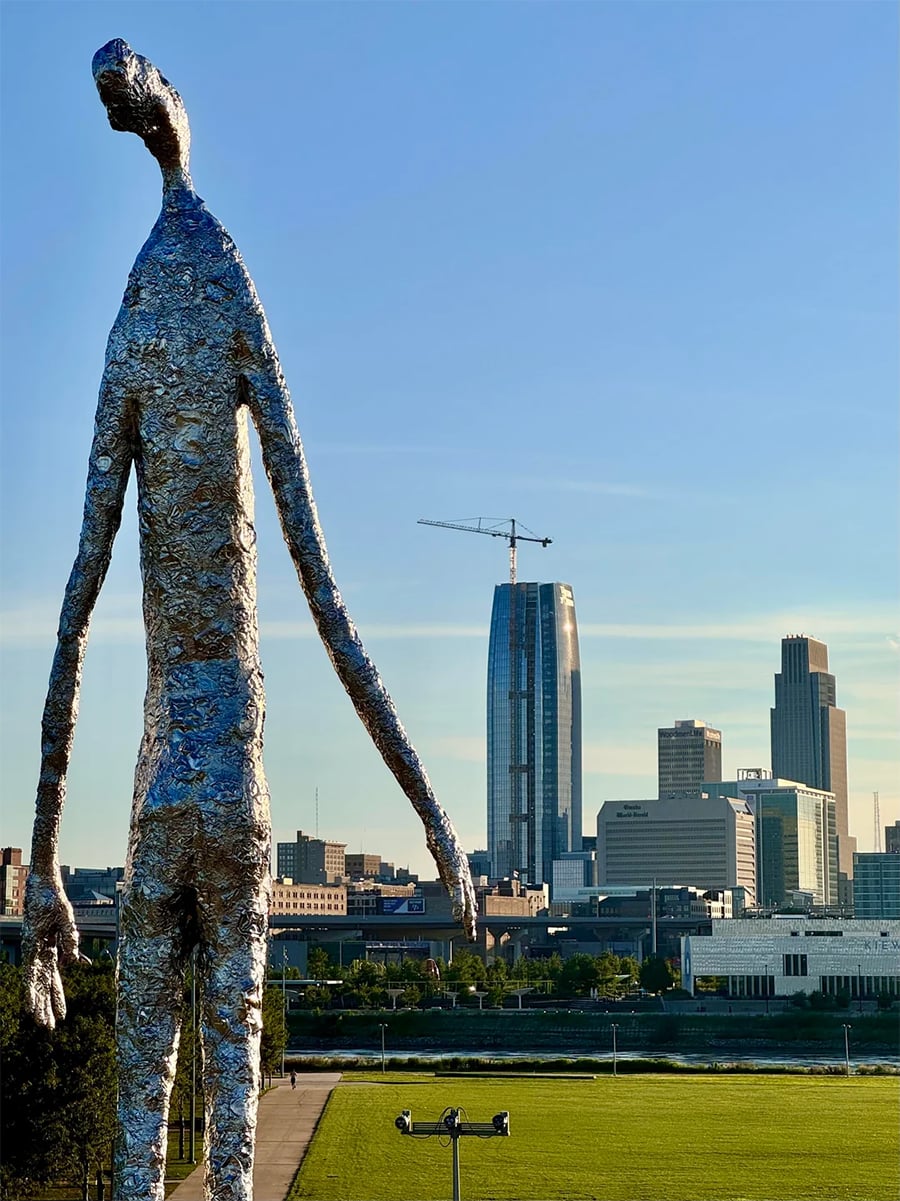 A tall, abstract silver sculpture stands in front of a city skyline with modern buildings, including a tall tower with a crane on top, under a clear blue sky.