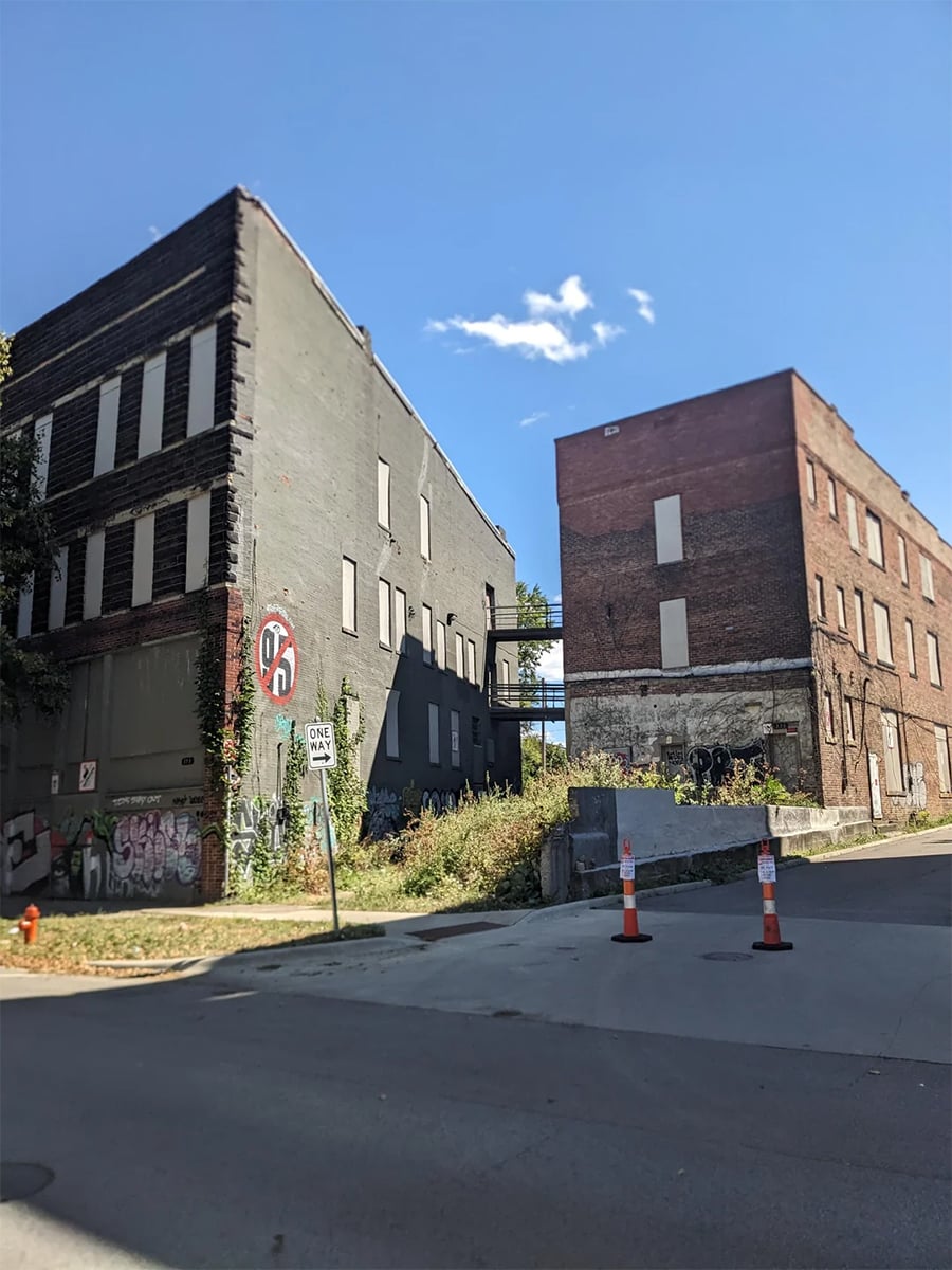 Two old, boarded-up brick buildings sit beside an overgrown lot. Graffiti covers the lower walls. Orange traffic cones and a no parking sign are visible on the street in front, with a blue sky and clouds overhead.