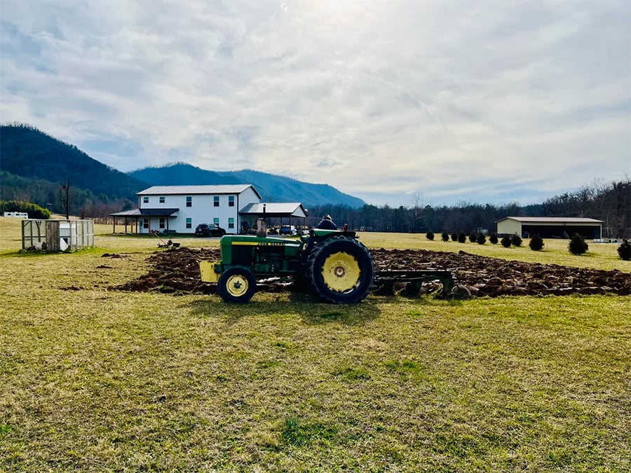 A green tractor plows a field on a grassy property with houses and mountains in the background under a cloudy sky.
