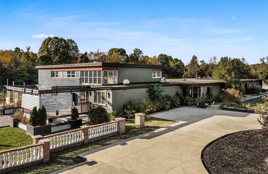 A modern, two-story house with large windows, a flat roof, a paved driveway, and landscaped yard surrounded by trees under a clear blue sky.