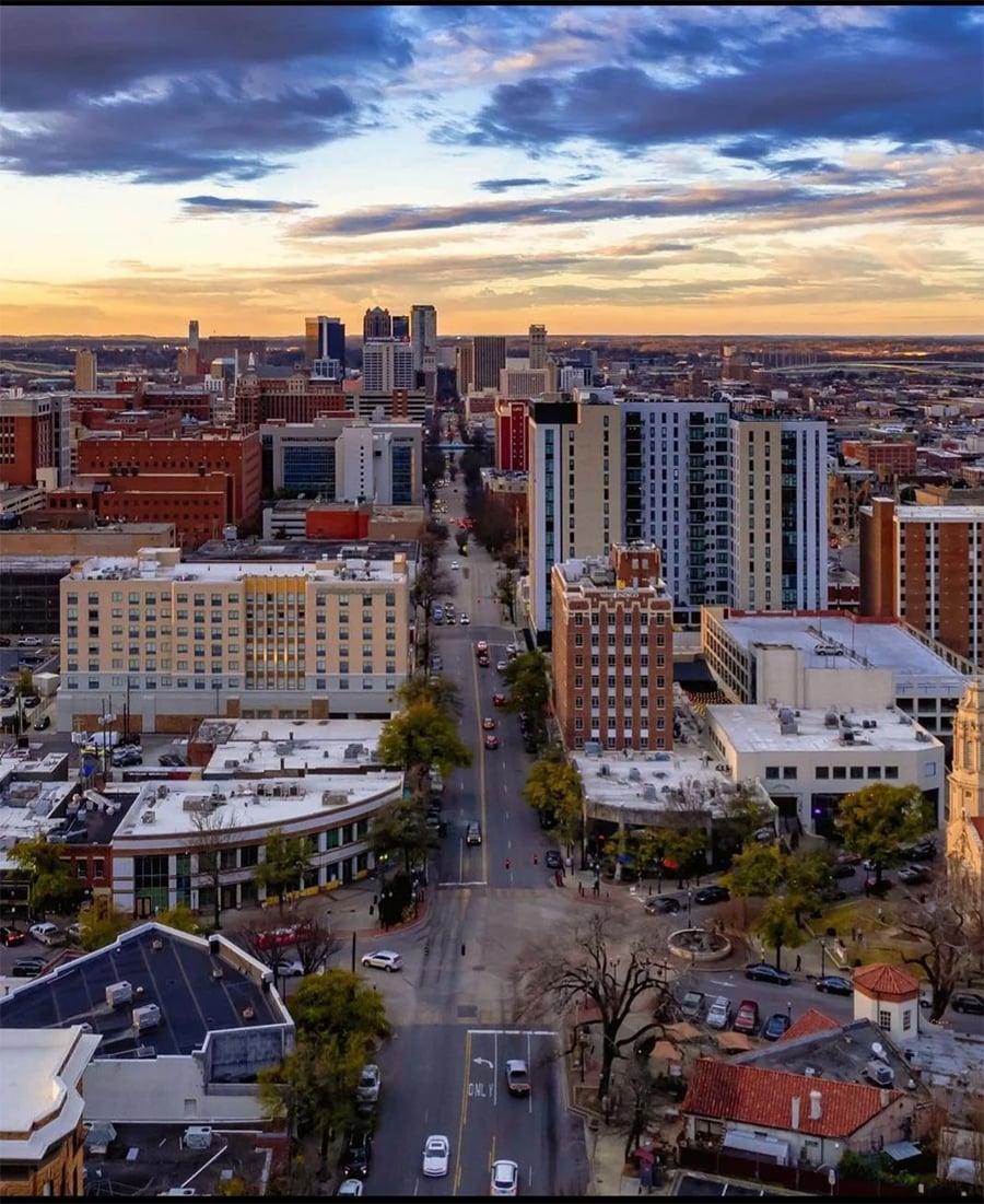 Aerial view of a cityscape at sunset, with tall buildings, streets lined with cars, and patches of greenery. A dramatic sky with clouds stretches across the horizon.
