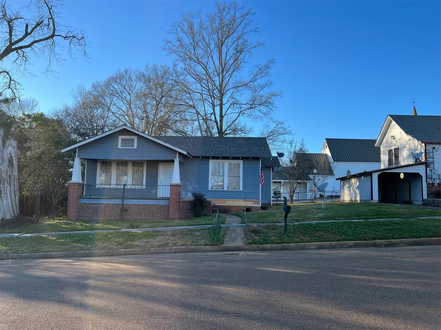 A small blue house with a front porch, brick foundation, and white trim; a flag hangs near the door. Trees and neighboring houses are in the background under a clear blue sky.