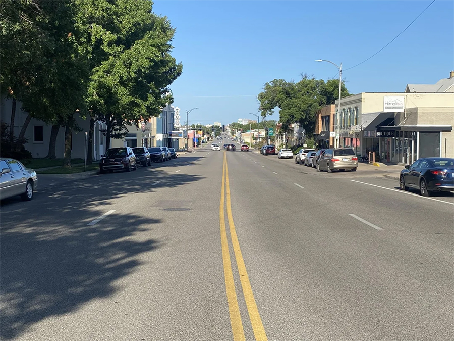 A wide, empty street with parked cars on both sides, buildings lining the road, and clear blue sky overhead. Trees provide shade on the left side of the street.