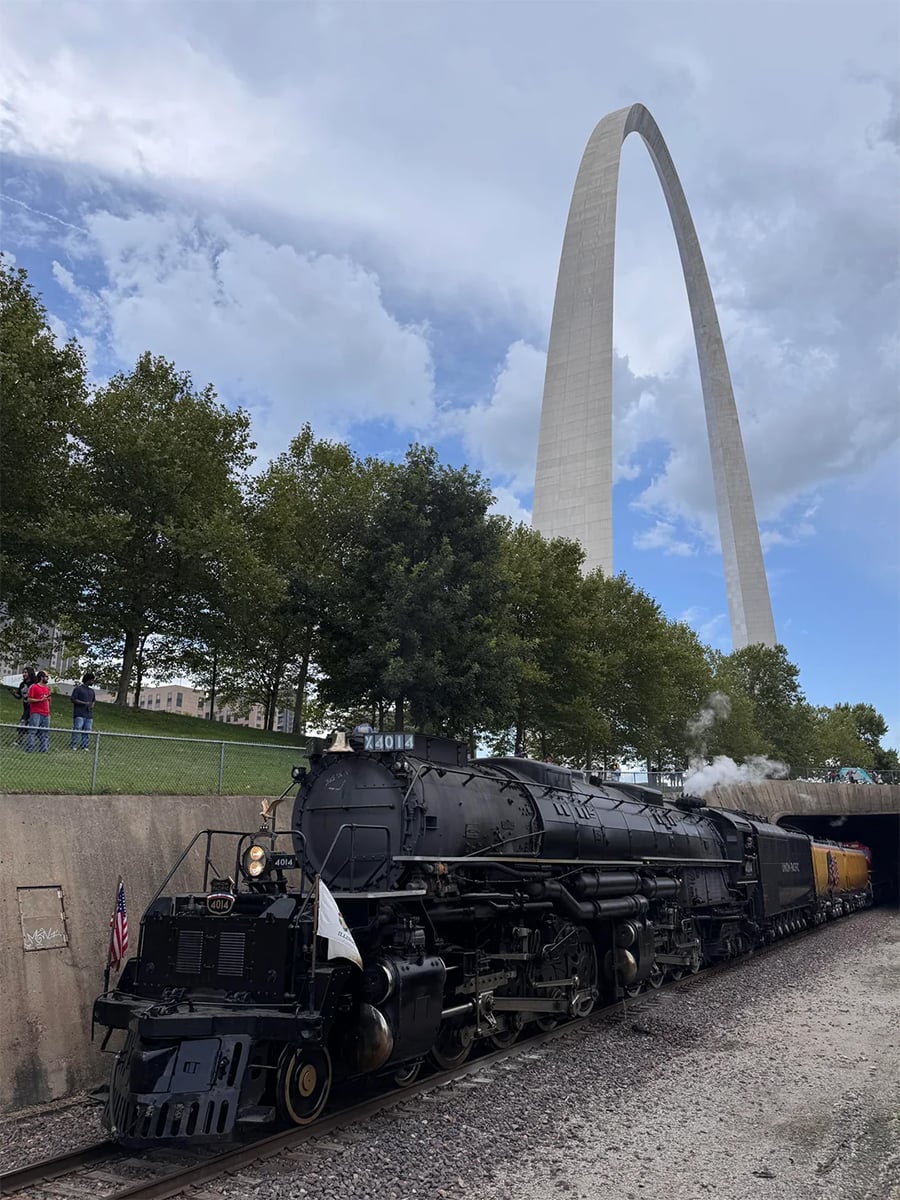 A black steam locomotive train passes near the Gateway Arch in St. Louis, with trees and clouds in the background and a few people standing on the left side, watching.